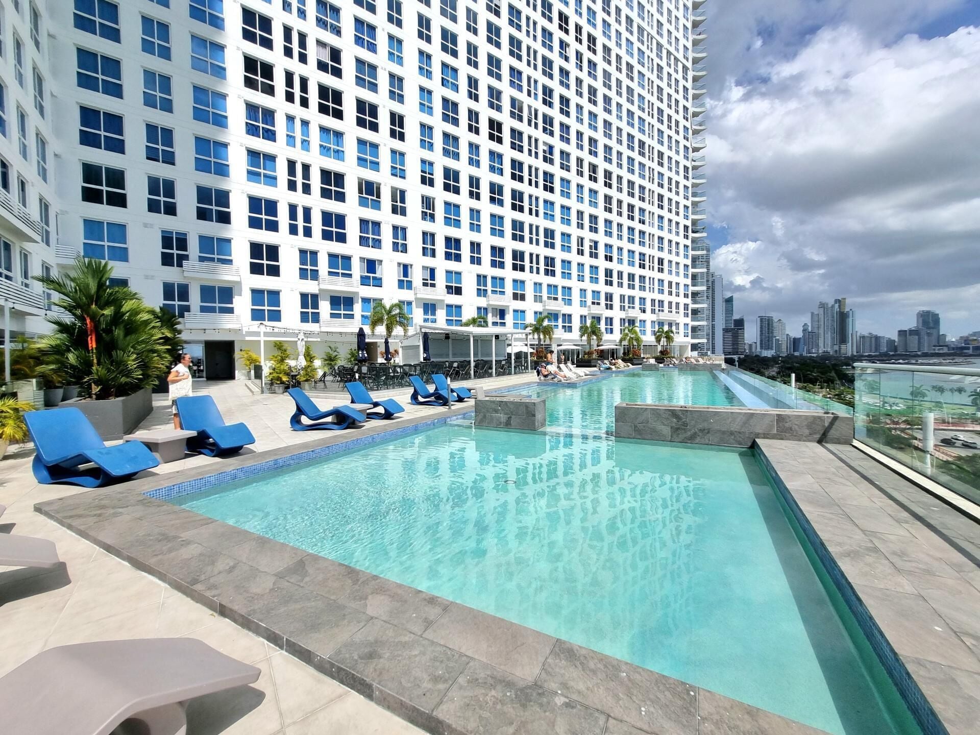 Un área de piscina en la azotea con sillas reclinables azules, una piscina azul cristalina y un horizonte urbano de fondo.