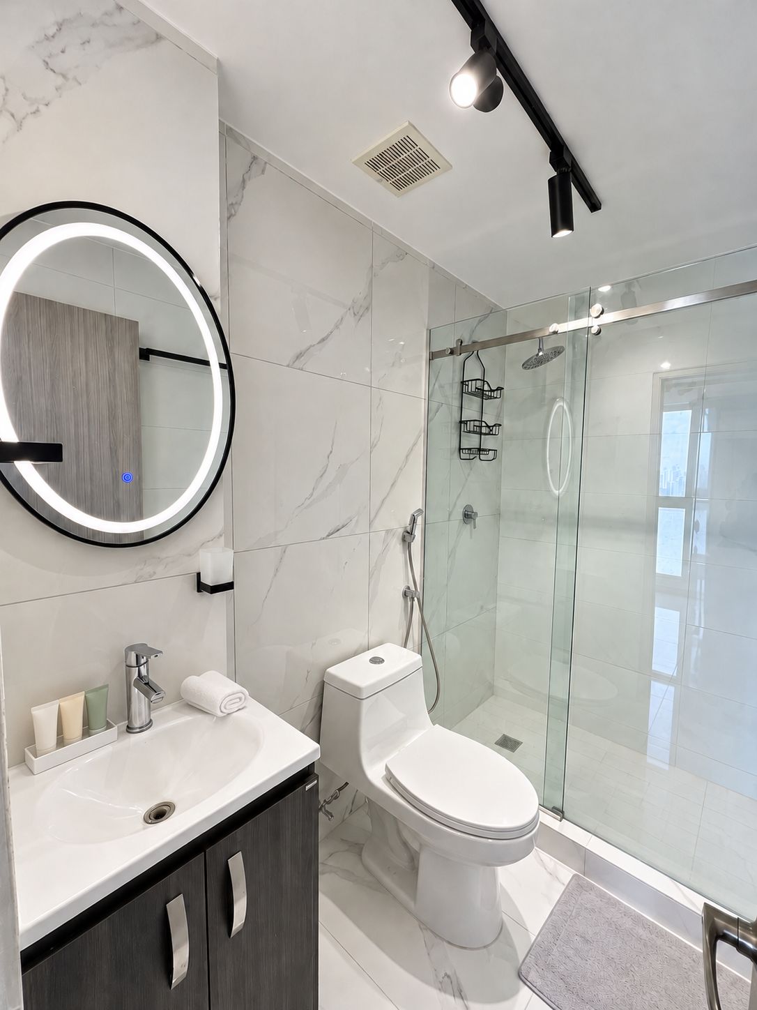 Een moderne badkamer met een marmeren spatwand, een ronde spiegel, een toilet, een douchecabine en een wastafel met een kastje.