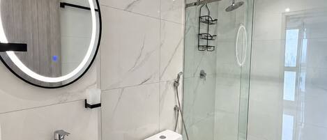 A modern bathroom with a marble backsplash, a round mirror, a toilet, a shower enclosure, and a vanity with a sink and cabinet.