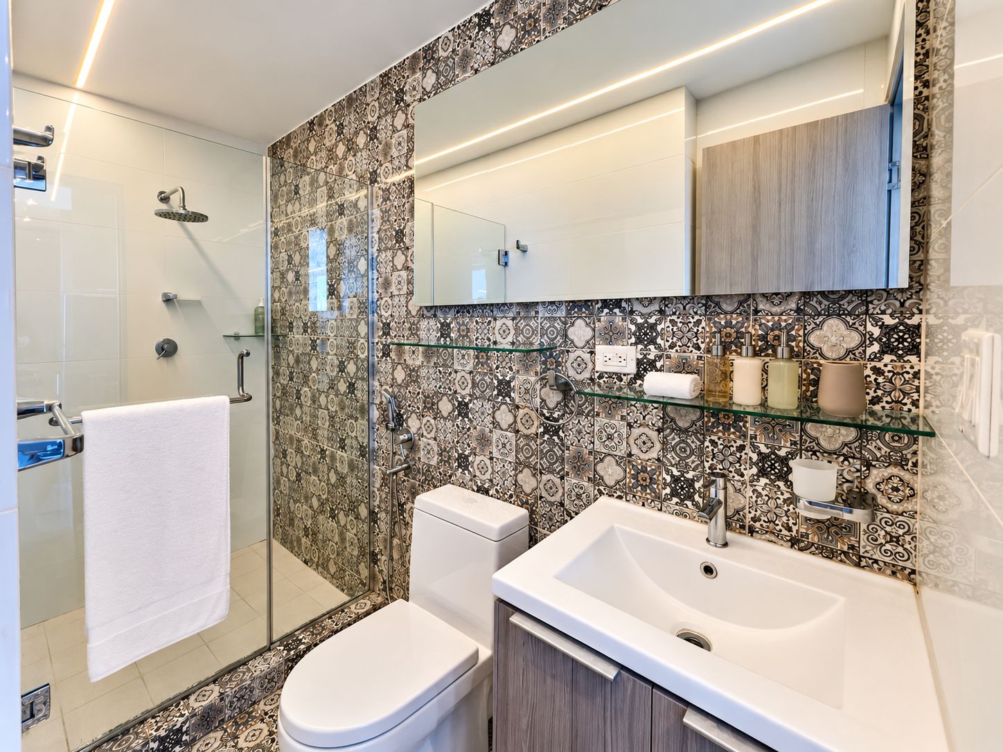 A modern bathroom with a glass shower enclosure, a toilet, a sink with a mirror above, and patterned tiles on the walls.