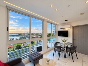 A modern living room with large windows, a flat-screen TV, and a dining area with chairs and a table. - Apartments at The Sands – Ocean View Panama City (Panama City)