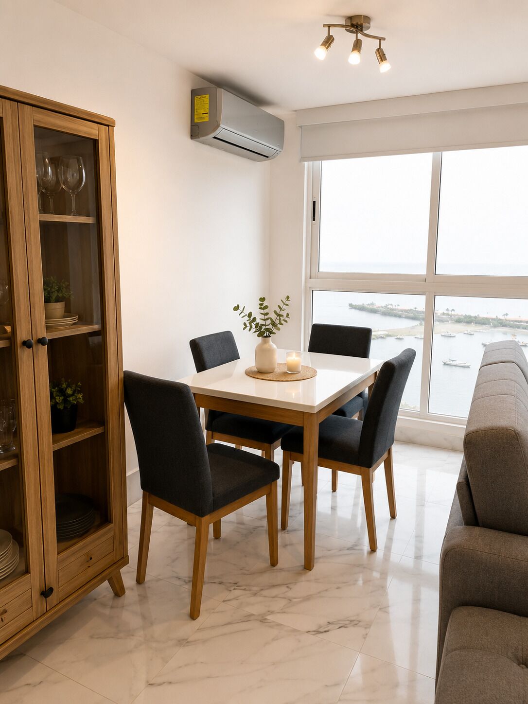 A dining area with a wooden cabinet, a table with chairs, and a view of the sea through large windows.