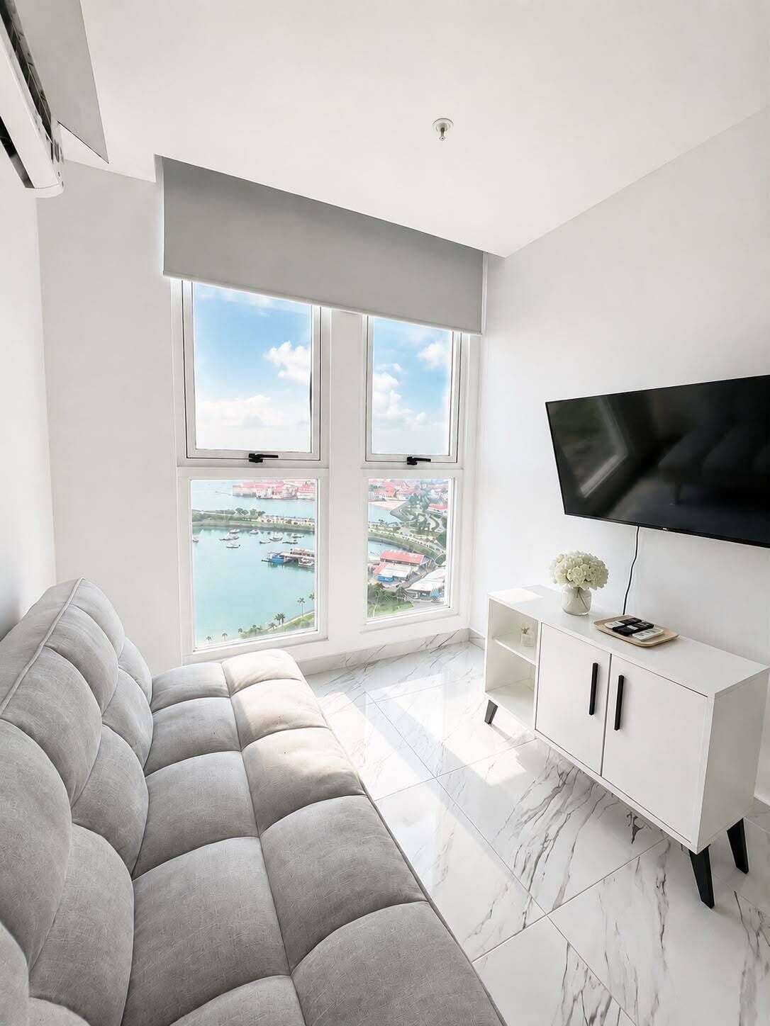 Una sala de estar moderna con un sofá, un televisor, un mueble blanco y ventanas amplias con vista al puerto.