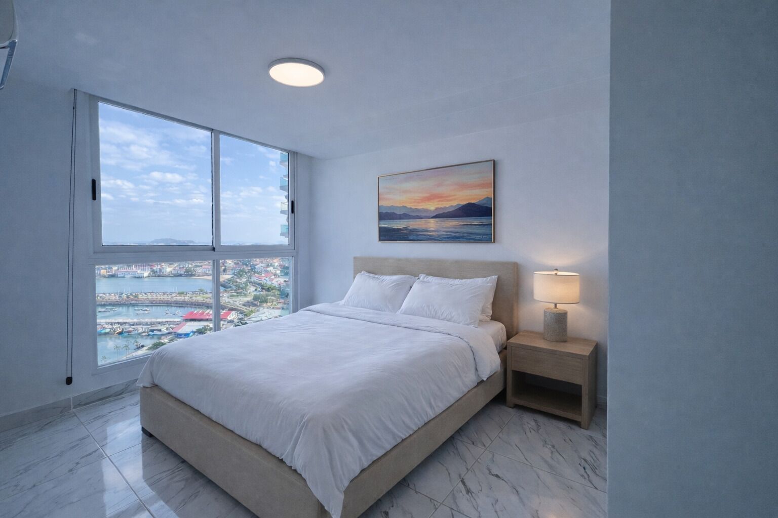 Une chambre à coucher comprenant un lit, une table de chevet, une lampe, un tableau et une grande fenêtre donnant sur la ville.