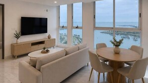 A modern living room with a large TV, a sofa, a dining table with chairs, and large windows with a view of the sea. - Apartments at The Sands – Ocean View Panama City (Panama City)