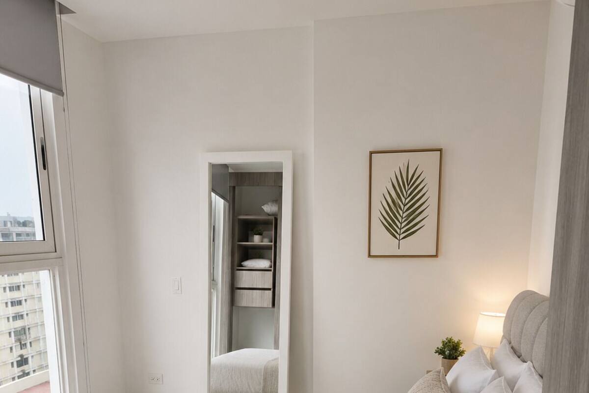 Habitación de hotel con cama, mesita de noche con lámpara, espejo, cuadro con una hoja y vista a la ciudad desde la ventana.