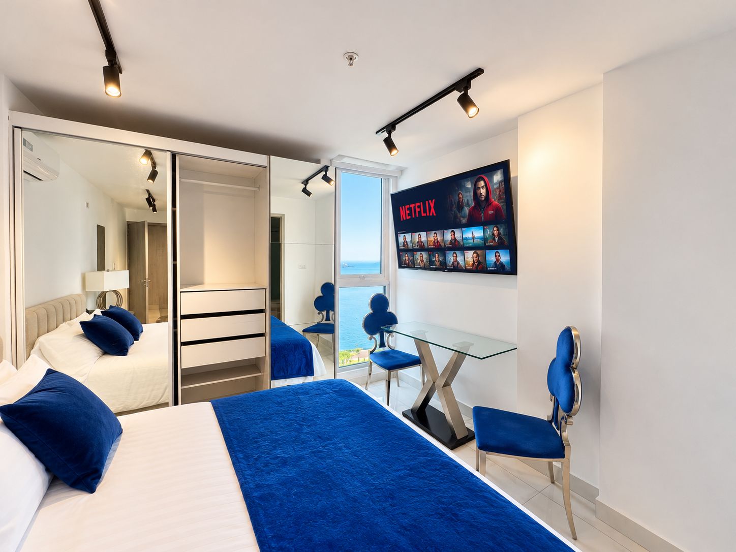 Quarto de hotel com cama, escrivaninha, cadeira, TV exibindo a interface da Netflix e varanda com vista.