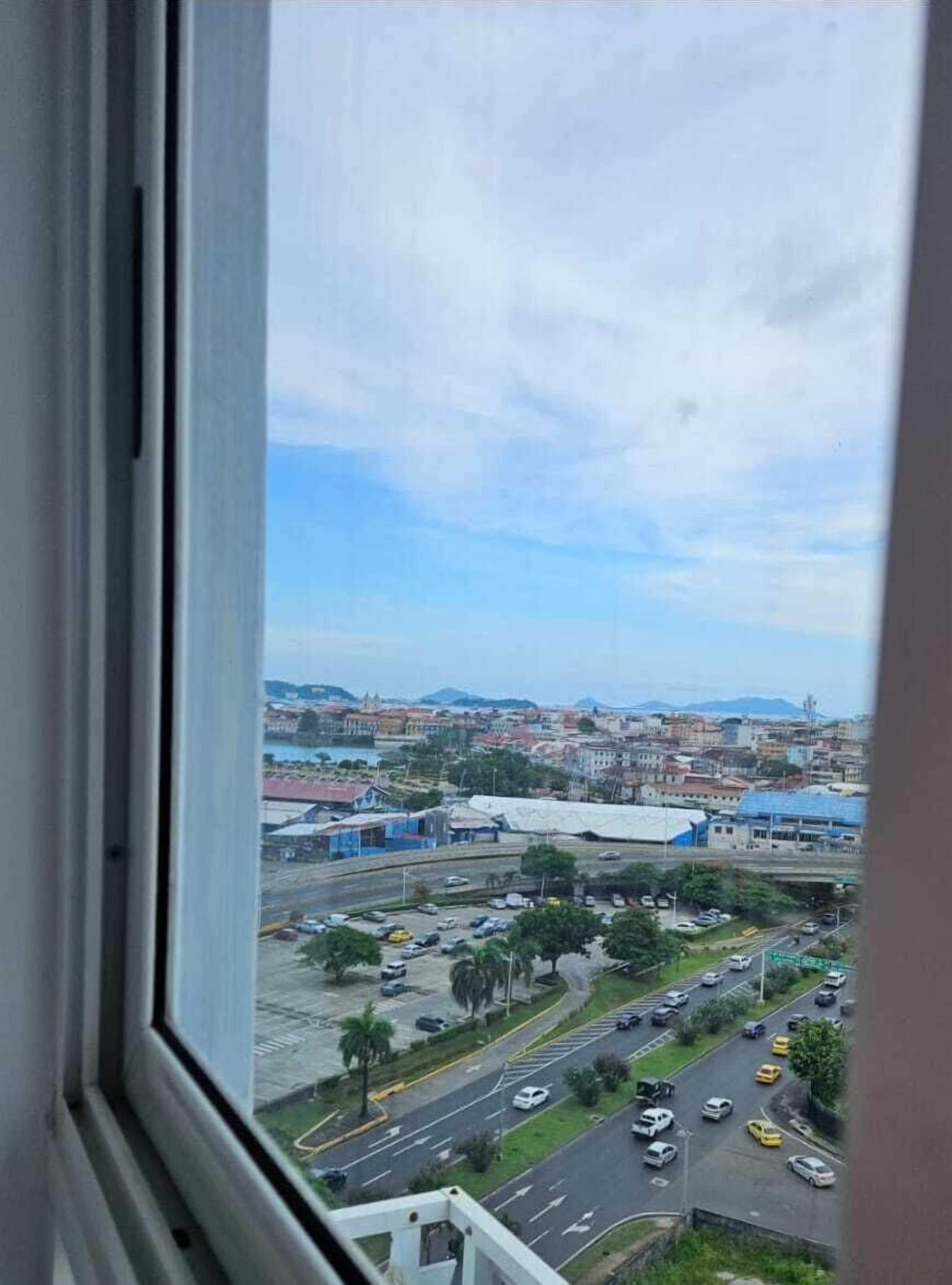 Un paisaje urbano a través de una ventana, con vista a una carretera, edificios y montañas distantes.