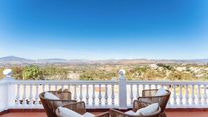 Ein Balkon mit Korbstühlen und einer weißen Brüstung, der auf eine Landschaft mit Bergen und spärlicher Vegetation blickt.