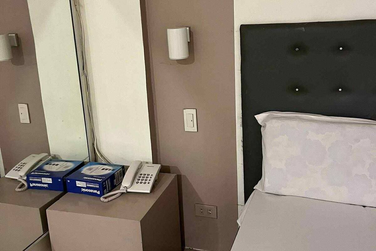 A hotel room with a bed, a large mirror, two bedside phones, and a small table with boxes.