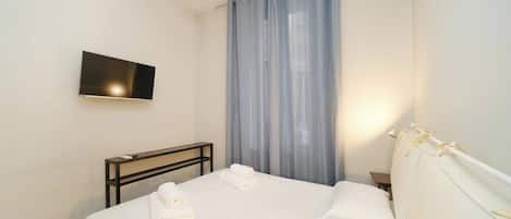 A hotel room with a bed, a flat-screen TV mounted on the wall, a bedside table, and a window with sheer curtains.