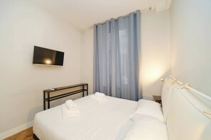 Ein Hotelzimmer mit einem Bett, einem flachbildfernseher an der Wand, einem Nachttisch und einem Fenster mit durchsichtigen Vorhängen.