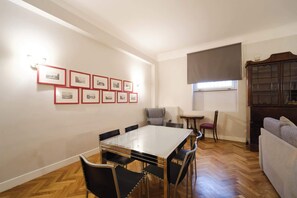 A dining area with a glass-top table and chairs, a radiator, a window with a blind, and framed pictures on the wall. - BuddyRent - Castel Sant'Angelo - 7 ospiti (Rome)