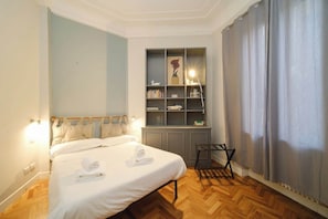 A bedroom with a bed, a bedside table, a shelf with books and decorative items, and a window with curtains.