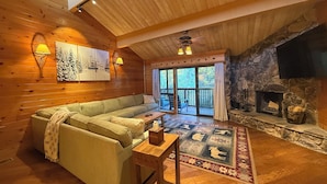 A cozy cabin interior with a wooden ceiling, a stone fireplace, a sofa, a small table, and a large window with a view of the outdoors.
