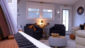 Una sala de estar con un piano, dos sillones, una mesa redonda y una puerta con un espejo y un reloj.