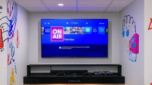 Una sala moderna con un televisor de pantalla plana grande montado en la pared, un mueble de entretenimiento negro debajo y dos sillas de juego negras con una alfombra colorida.