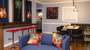 Una sala de estar con un sofá azul, cojines coloridos, un taburete rojo, una zona de comedor con una mesa y sillas, y una pared decorada con guitarras enmarcadas y un cuadro abstracto lleno de color.
