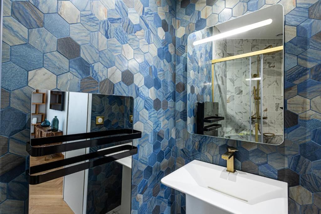 A modern bathroom with a white sink, gold faucet, and a mirror above it. The walls are covered with blue and gray hexagonal tiles.