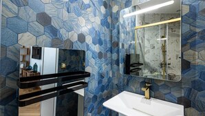 A modern bathroom with a white sink, gold faucet, and a mirror above it. The walls are covered with blue and gray hexagonal tiles.