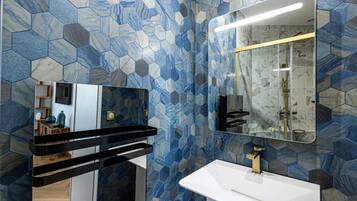 A modern bathroom with a white sink, gold faucet, and a mirror above it. The walls are covered with blue and gray hexagonal tiles.