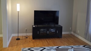 A room with a flat-screen TV on a stand, a floor lamp, and a built-in air conditioning unit on the wall. - The Grand Estate- 4 Bedroom  (Moncton)