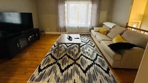 A living room with a sofa, a coffee table, a TV, and a large window with curtains. - The Grand Estate- 4 Bedroom  (Moncton)