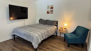 Een slaapkamer met een bed, een blauwe fauteuil, een nachtkastje met een lamp en een aan de muur bevestigde televisie.