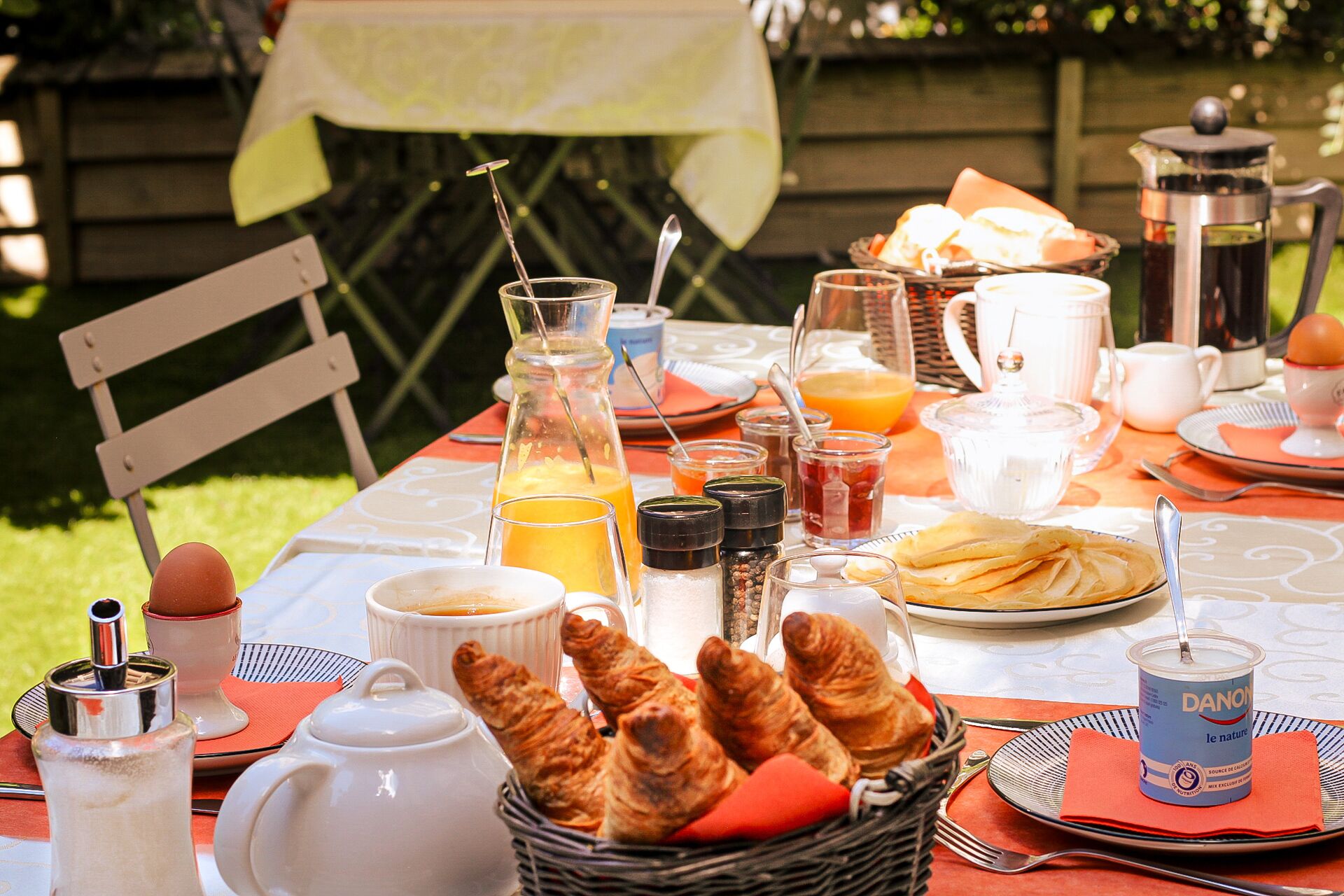 A breakfast table set outdoors with croissants, coffee, orange juice, and a variety of condiments.
