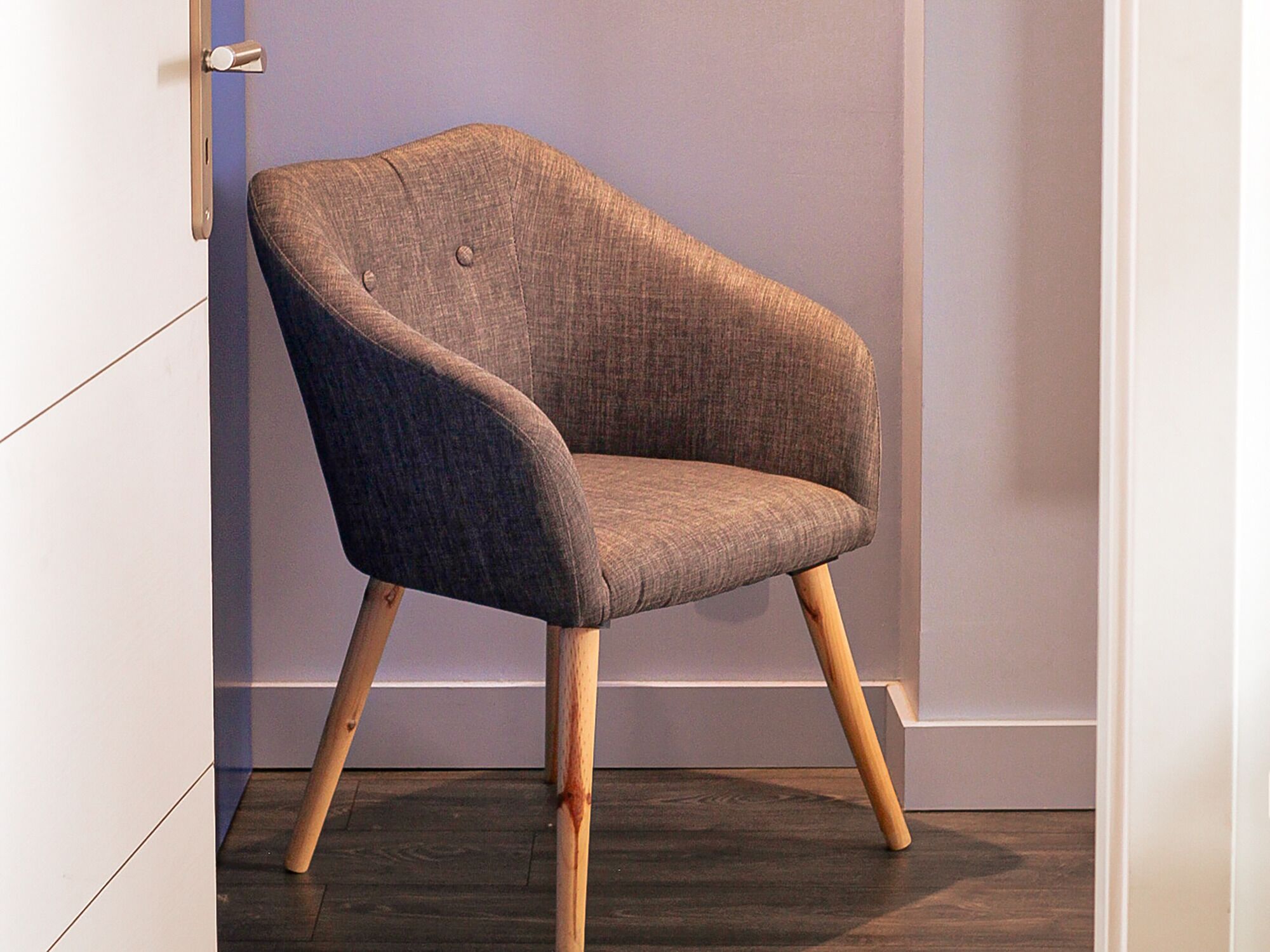 Een moderne fauteuil met houten poten en een gestructureerde stof, geplaatst in een kamer met een effen muur en een deur.