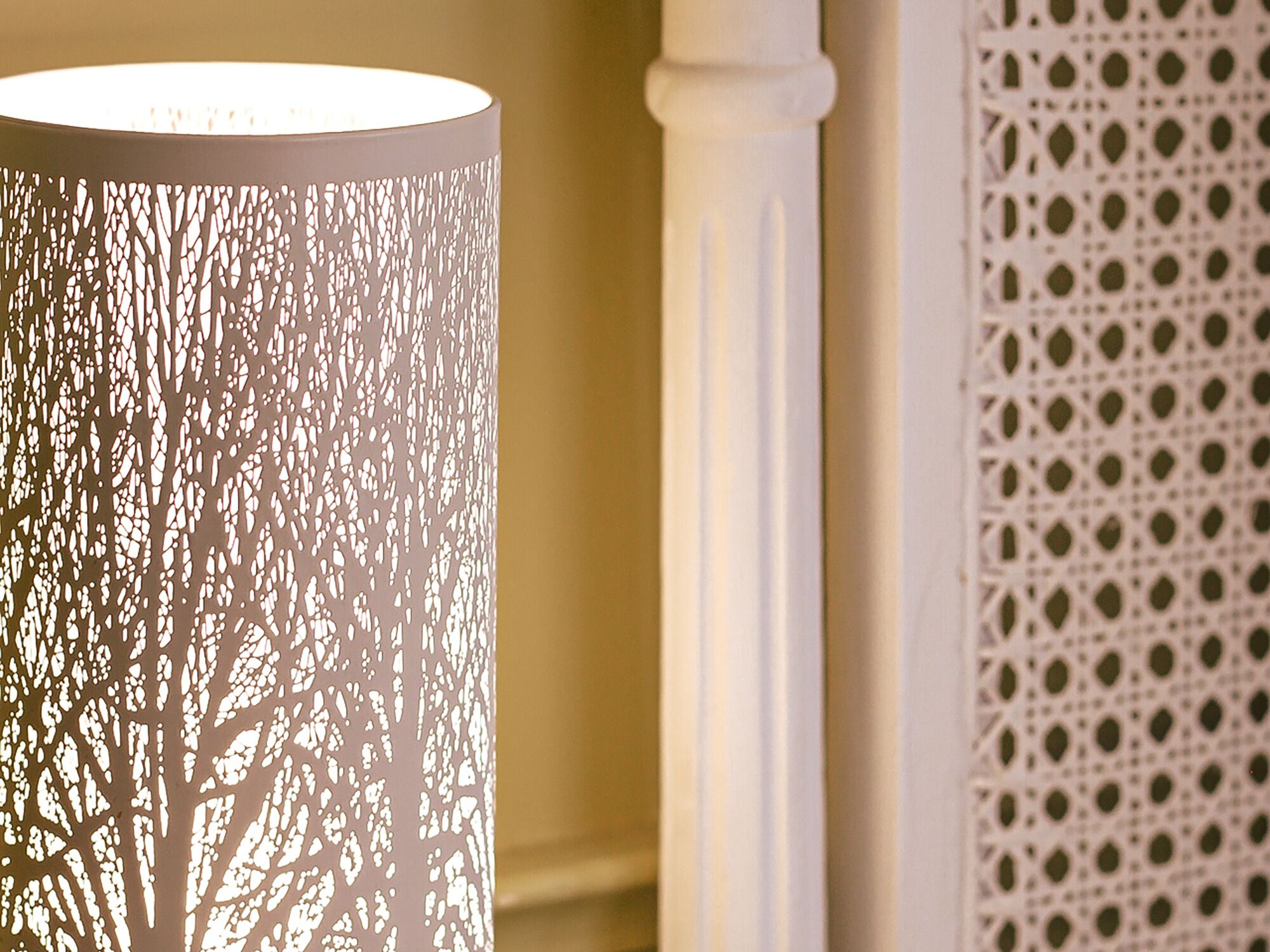 En cylindrisk lampa med ett träliknande, perforerat mönster, en vit pelare och en perforerad väggpanel.