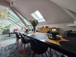 Un comedor moderno estilo loft, con techo alto y tragaluz, que presenta una mesa de madera, sillas negras y un jarrón con flores.