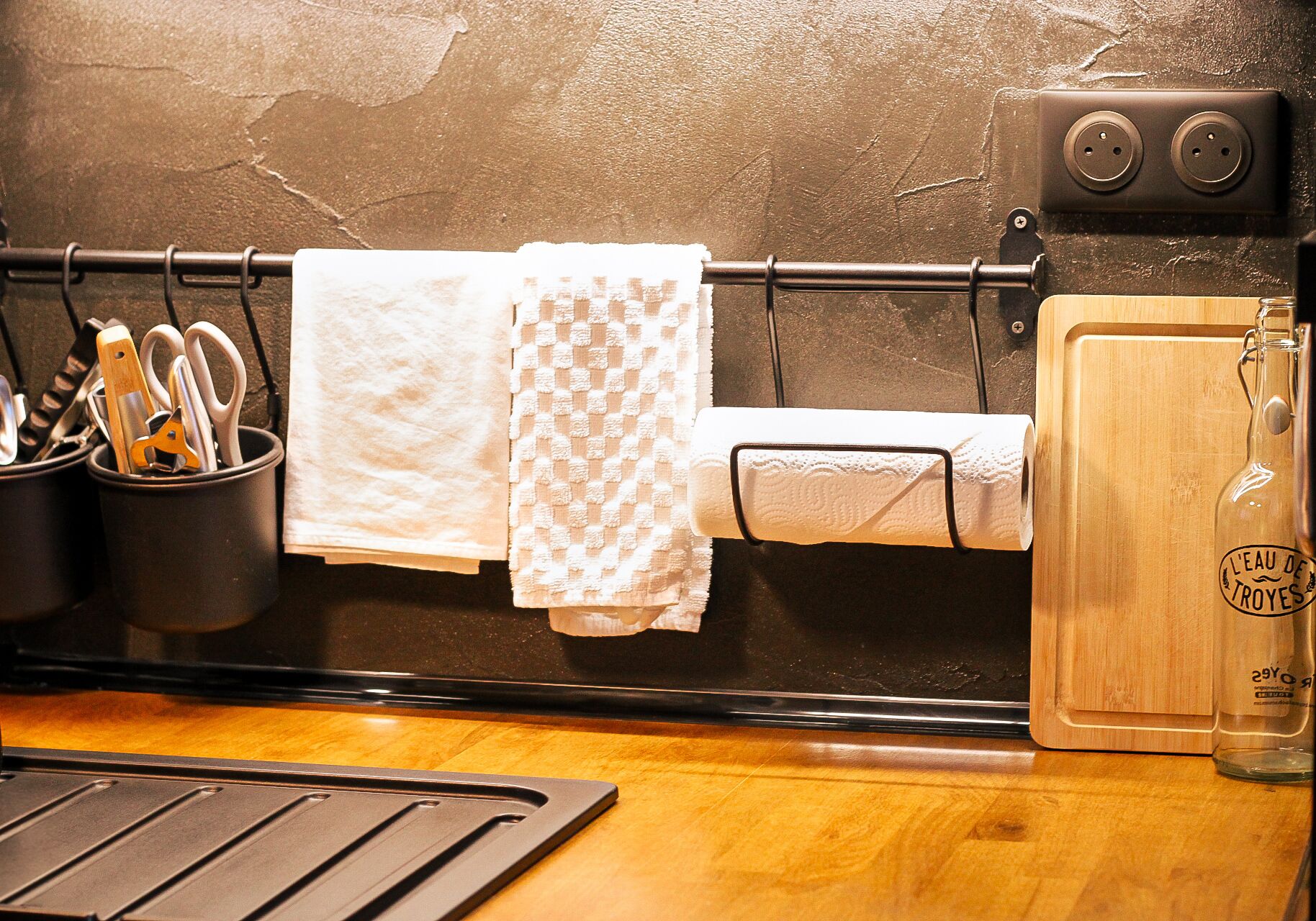 A kitchen with a black countertop, a cutting board, a bottle of water, and a towel rack with towels.