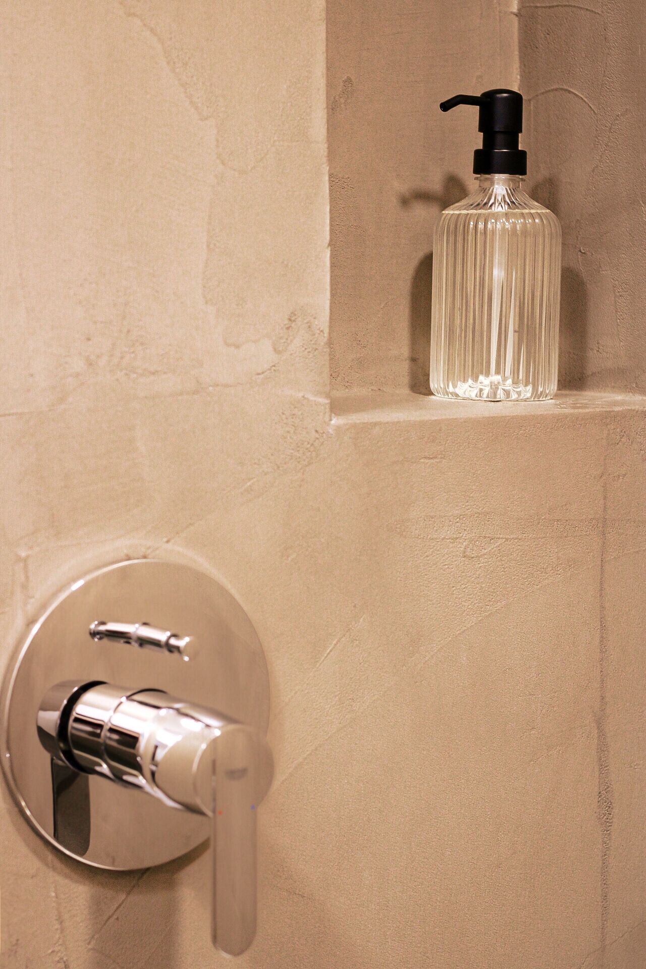A bathroom with a round, chrome-finished faucet and a clear glass bottle with a black pump dispenser on a beige wall.