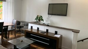 A modern living room with a flat-screen TV mounted on the wall, a wooden coffee table, a glass dining table with chairs, and a potted plant.