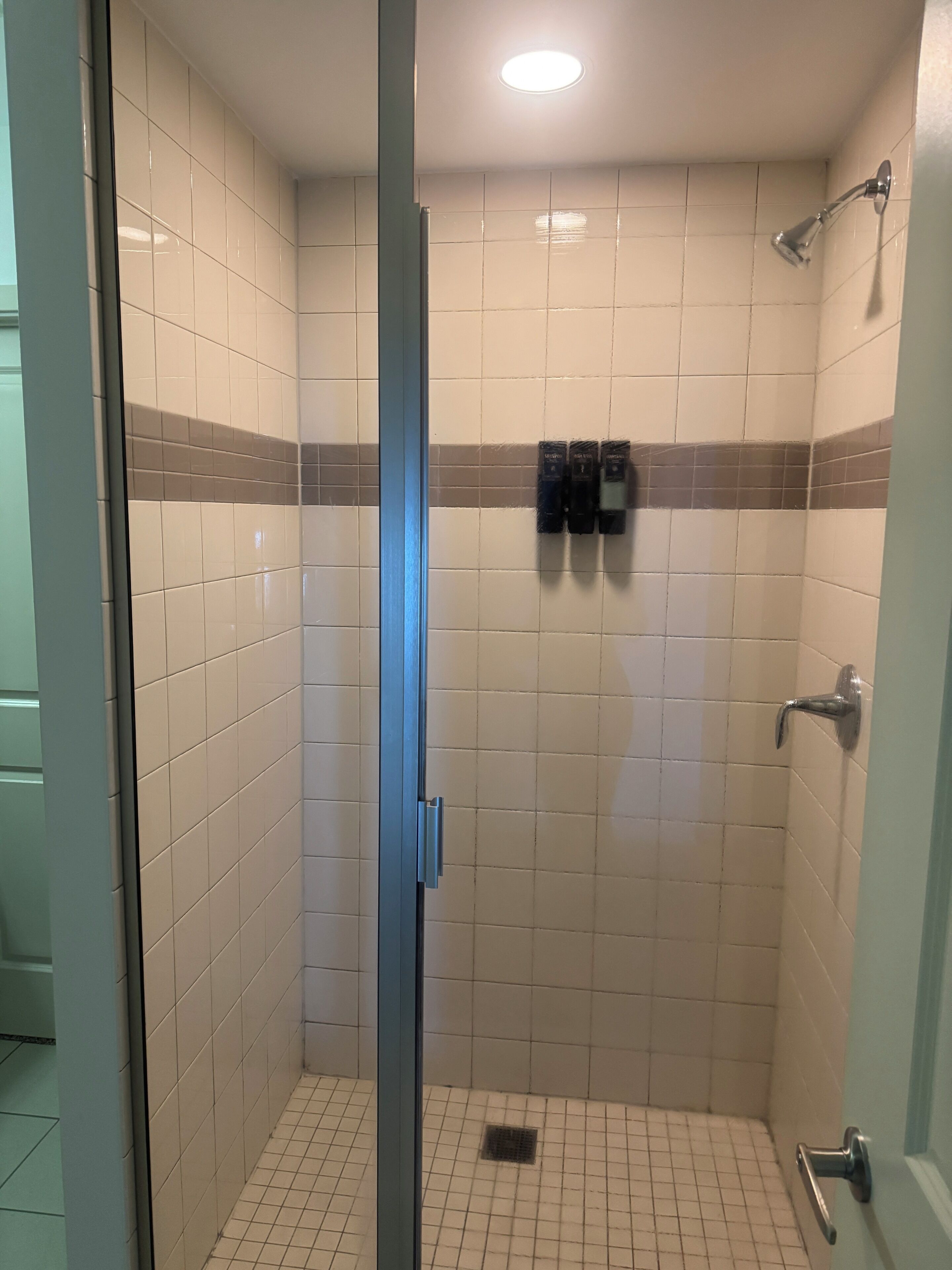Een kleine douchecel met betegelde wanden, een glazen deur, douchekop en een handdoekrek met toiletartikelen.