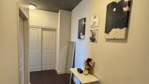 A hallway with dark wooden flooring, white walls, and a ceiling light. There are three framed artworks on the wall and a small white table with a vase and candles.