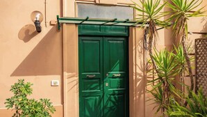 Eine grüne Tür mit einem kleinen Fenster darüber, einer Wandleuchte und einer Pflanze zur Rechten.