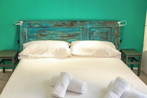 Een bed met witte lakens, twee kussens en twee opgerolde handdoeken tegen een muur in teal.