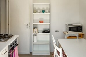 A compact kitchen with a built-in oven, a microwave on a small table, a white cabinet with shelves, and a pink towel hanging on the oven door.