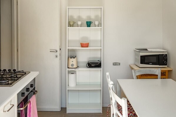 Une cuisine compacte avec un four encastré, un micro-ondes posé sur une petite table, un meuble blanc avec des étagères, et une serviette rose accrochée à la porte du four.