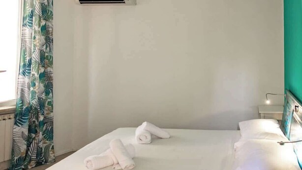 Ein ordentlich bezogenes Bett mit weißen Leinen und aufgerollten Handtüchern, ein Nachttisch mit Lampe und ein Fenster mit Vorhängen.