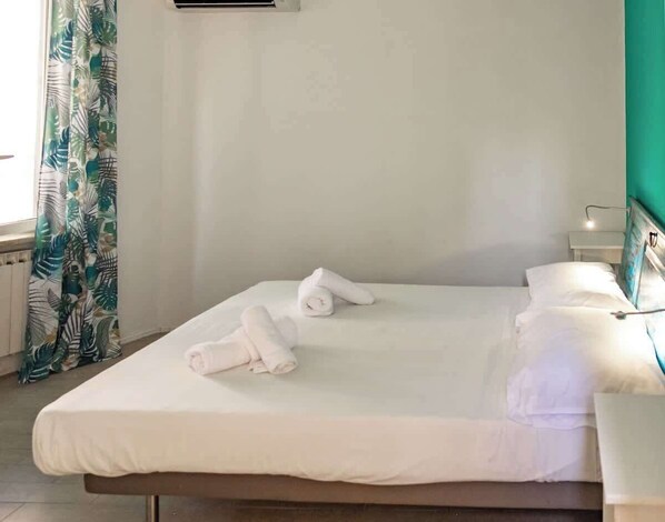 Een net opgemaakt bed met witte lakens en opgerolde handdoeken, een nachtkastje met een lamp, en een raam met gordijnen.