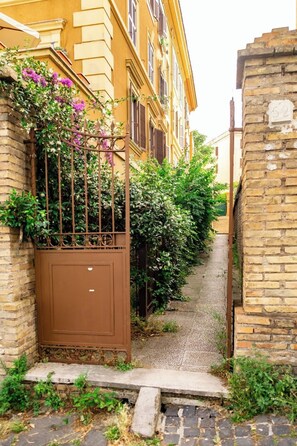 Une ruelle étroite, avec un portail en métal, un mur de briques et un immeuble jaune aux volets fermés.