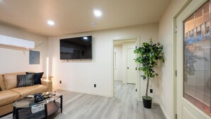 A modern living room with a beige sofa, a glass coffee table, and a flat-screen TV mounted on the wall. - Modern Cabin  Mins from TU, Downtown, Expo, St John and HIlcrest - upstairs (Tulsa)