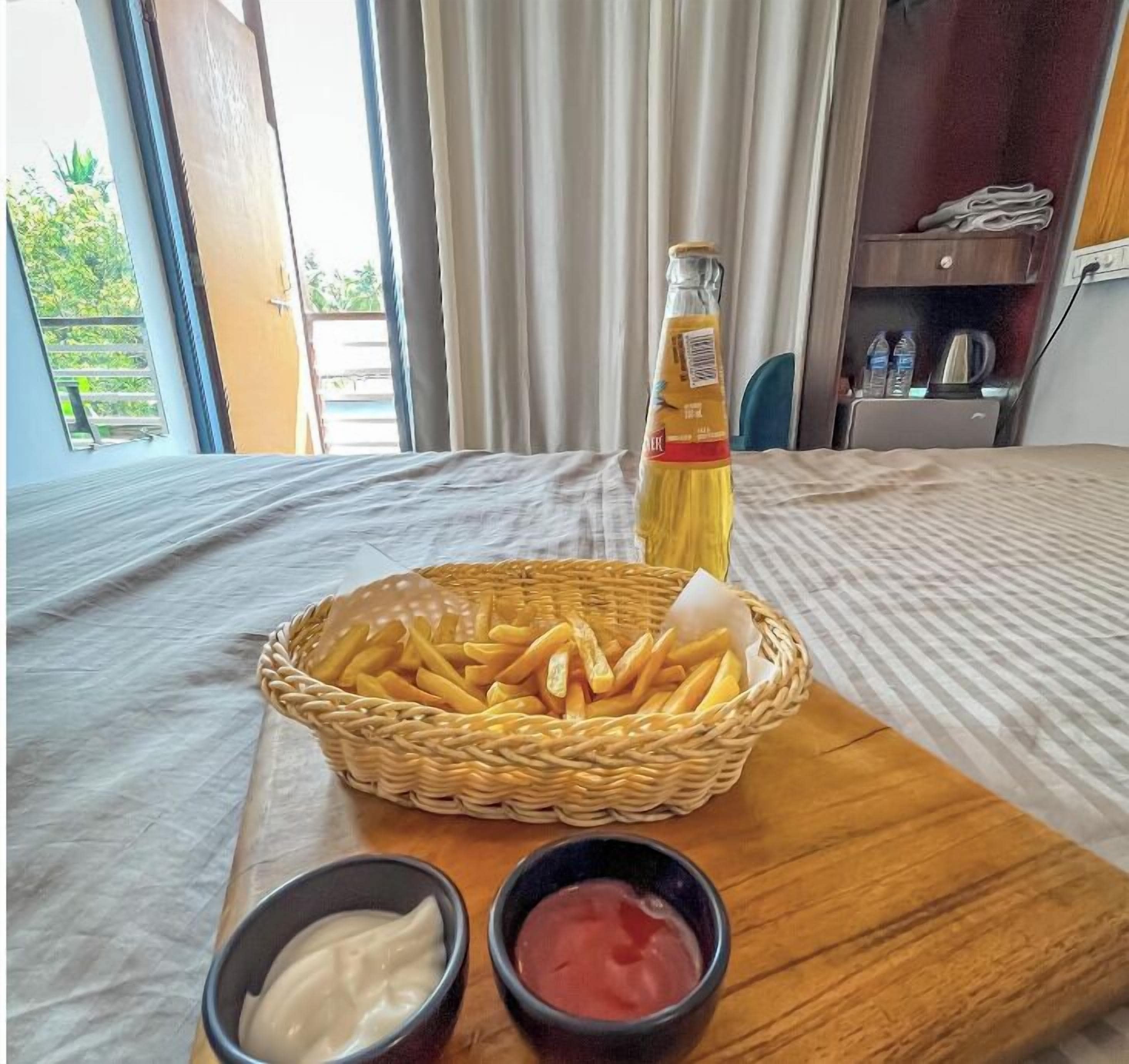 Una cama con cabecera de madera, una cesta de mimbre con papas fritas, una botella de salsa y dos recipientes pequeños con acompañamientos sobre una bandeja de madera.