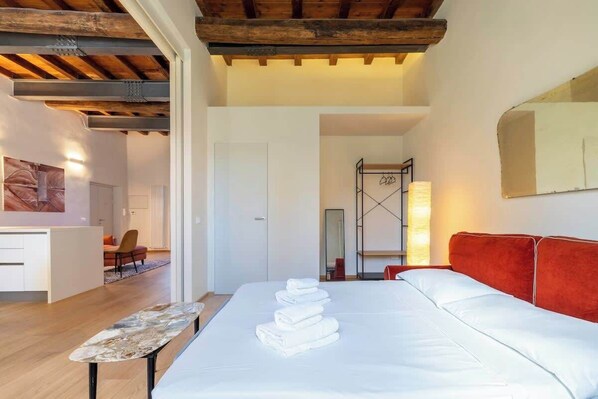 Habitación de hotel con cama, una mesita, sofá rojo, espejo y baño al fondo.