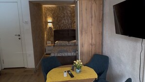 A compact living space with a dining area featuring a round table and four chairs, a bed with a headboard, a television mounted on the wall, and a door leading to another room.