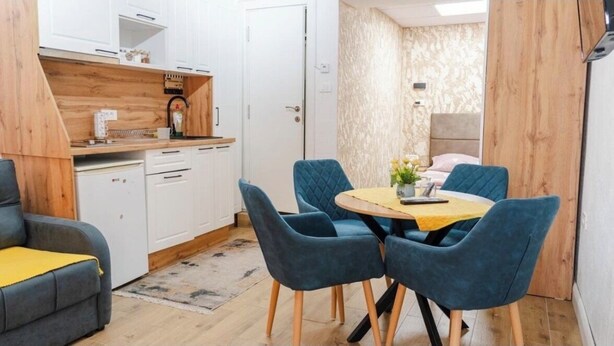 A compact living space with a kitchenette, a dining area with blue chairs, and a bed in the background.