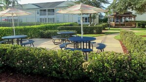 Ein Terrassenbereich im Freien mit mehreren Tischen und gestreiften Sonnenschirmen, umgeben von gepflegten Hecken und einem Gazebo.